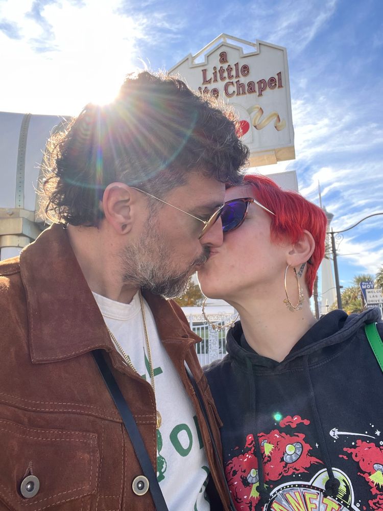 A couple shares an intimate moment in front of the "Little Chapel" sign in Las Vegas. The person on the left wears a brown leather jacket over a white shirt, while their partner on the right has vibrant red hair, sunglasses, and a black graphic hoodie. The sun creates a lens flare in the upper portion of the image, with a bright blue sky and white clouds in the background.