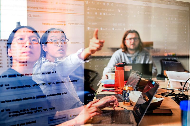 Three researchers collaborating at a previous ImmunoX Computational Biology Initiative (ICBI) Hackathon. They are gathered around a computer, looking at a dataset on the screen while one points at it.
