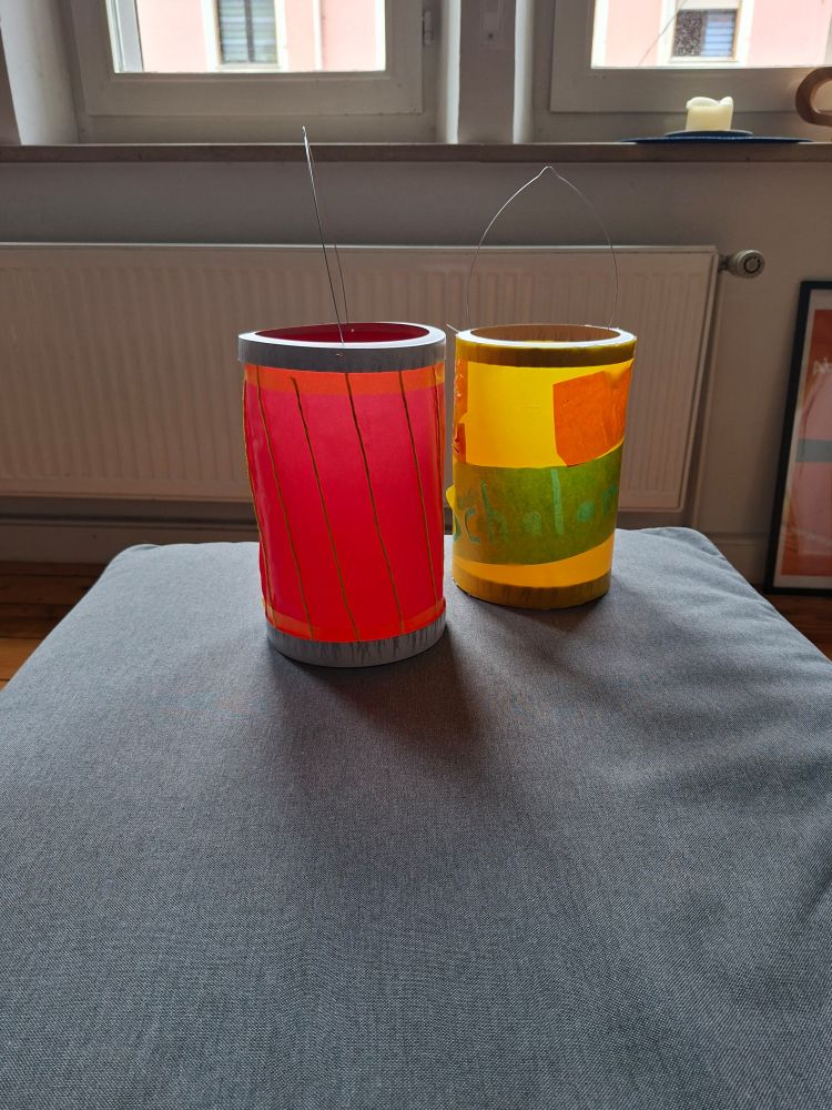 Zwei Laternen in klassischer Zylinderform, die eine ist rot mit gelben längsstreifen und die andere gelb mit grün und orangen patches.
