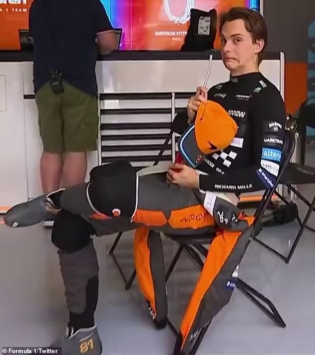 oscar piastri sitting in a chair with his left leg crossed over his right, he is holding his hat. oscar is looking at the camera with a face that gives the same vibes as “uh oh, that’s awkward” 
