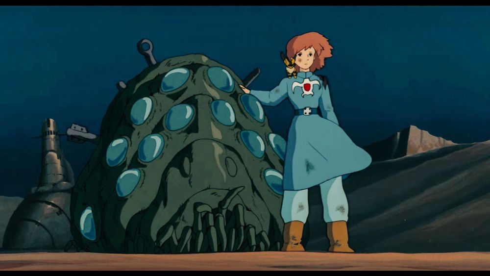 Cena do Anime japonês chamado Nausicaa, mostrando a personagem de mesmo nome, uma garota de cabelo curto castanho com um sobretudo azul ao lado de um monstro que parece um tipo de crustáceo gigante como um rinoceronte com dezenas de olhos azuis no formato de um tipo de verme com uma carapaça similar a uma lagosta.