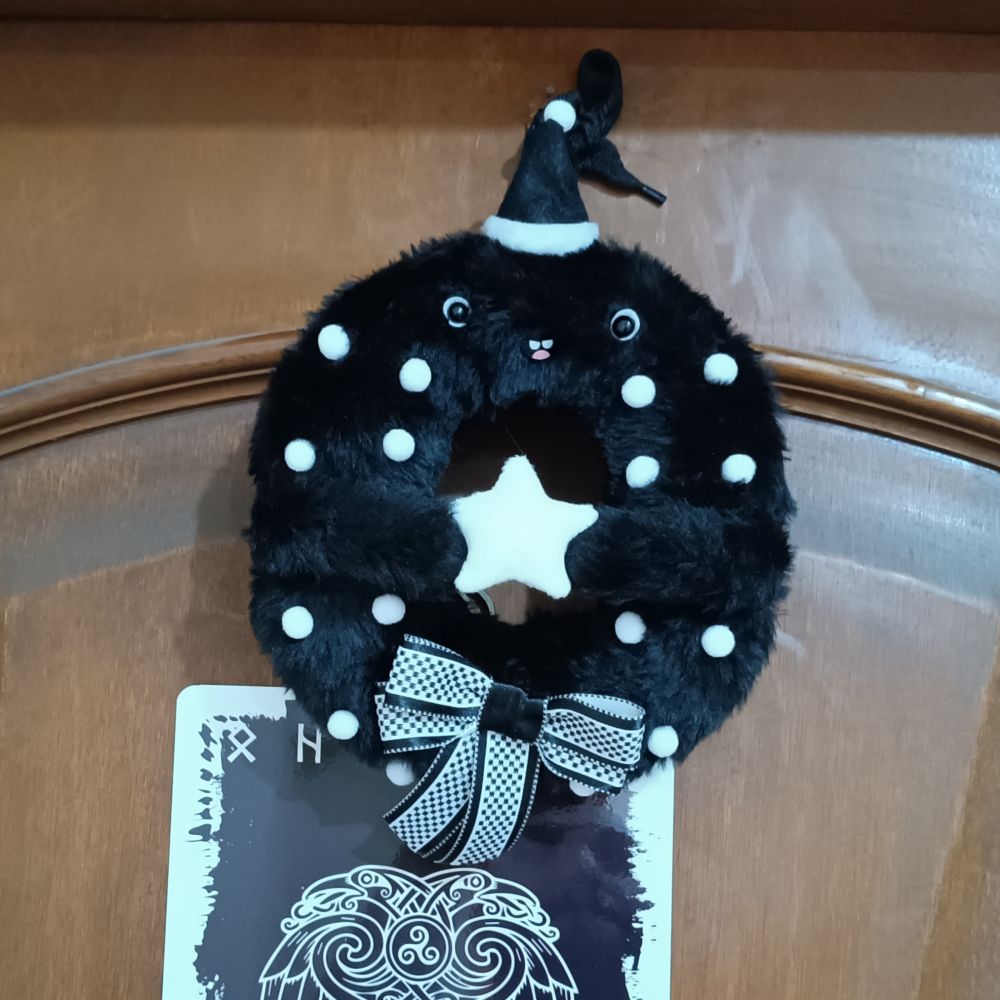 Foto da parte superior de uma porta marrom com um relevo de arco na madeira adornado com um friso elevado com uma guirlanda preta e branca feita de pelúcia com dois olhos como de desenho animado e uma pequena boquinha com dois dentinhos e uma língua rosinha numa expressão alegre e fofa, usando também um gorro de Natal preto e branco e enfeitada com bolinhas brancas e com dois bracinhos segurando uma estrela branca. Abaixo na porta se vê uma placa preta e branca com tunas nórdicas e um padrão viking de desenho de dois corvos entrelaçados. 