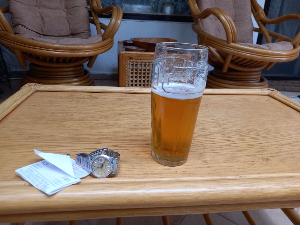 A bamboo table, outside on the terrace, with a beer glass and a watch sitting on a receipt.

I shall do no more today.