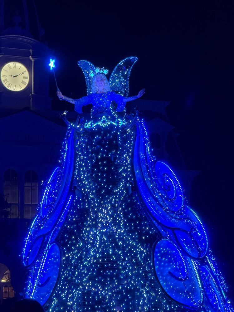 A woman wearing a lighted crown and holding a wand. She has  lighted wings. Her gown is made up entirely of blue lights. 