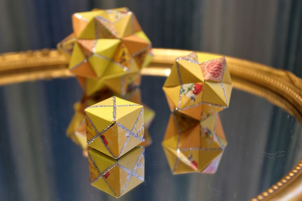 Photograph of a set of three different sized Kusudama. They have been placed on an old mirror that has a gilded oblong frame. The background is my trusted turquoise velvet curtains. The Kusudama are all yellow with panted details in glitter. 