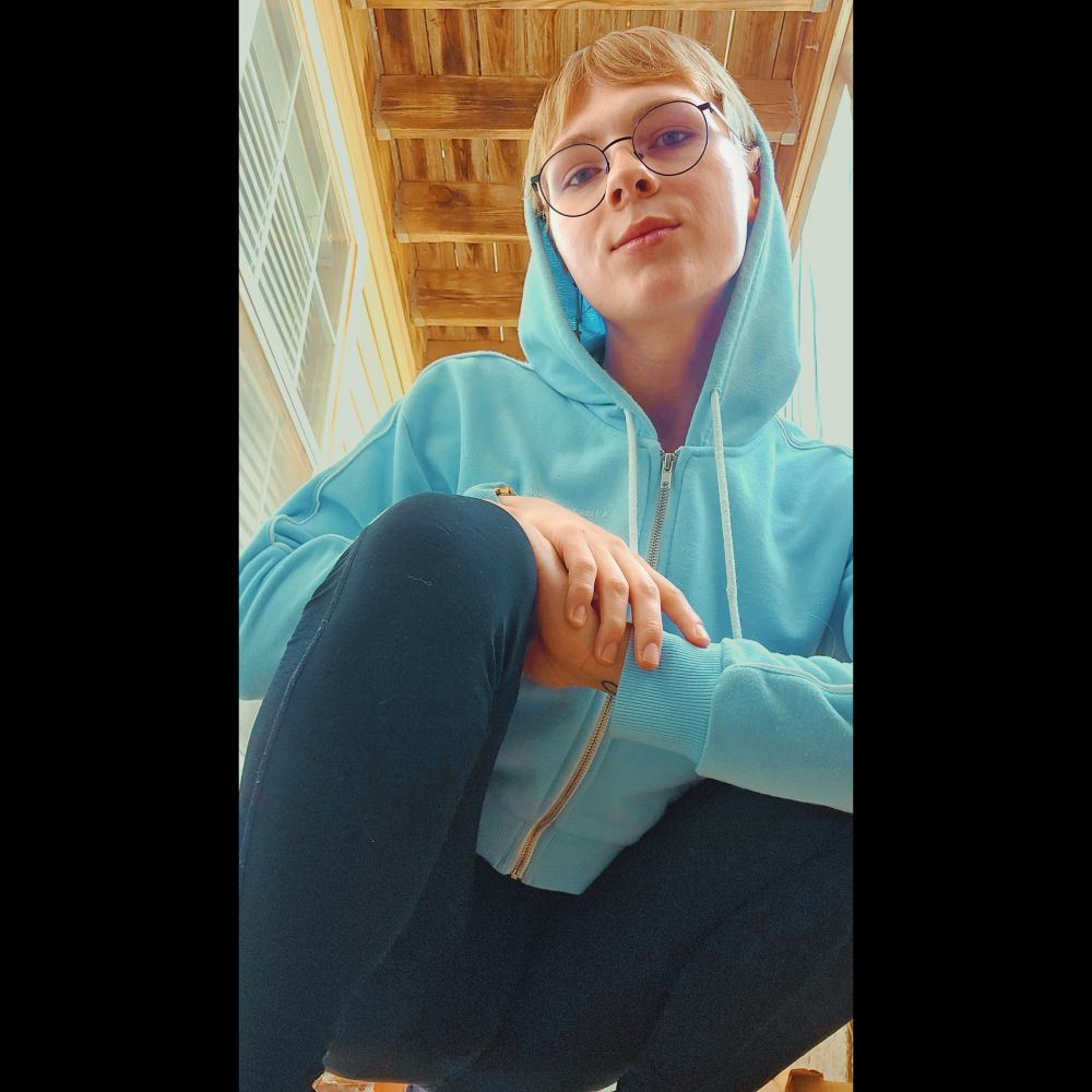 Renna (me!!!), a blonde, blue-eyed girl with glasses, squatting at a slight angle, looking towards the camera with her hands clasped on her knee

she is wearing a light blue cropped hoodie and black leggings