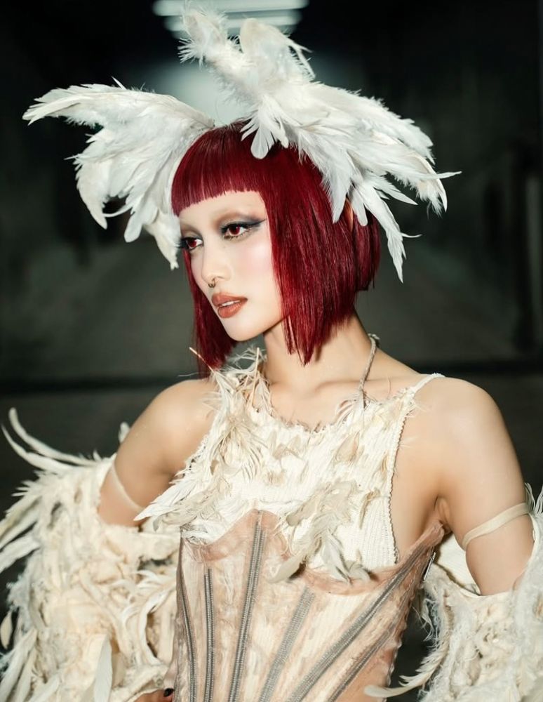 close up picture of jurin from xg in a feathered gown, with short red hair with bangs and a smoky eye shadow look 