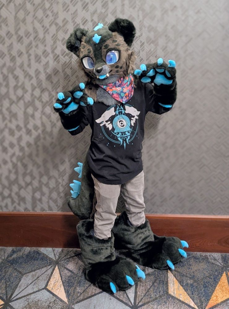 A person wearing a partial fursuit of a Godzilla-inspired dog standing in a hotel hallway. The character is dark gray with spotty markings, floppy ears, and bright blue accents. 