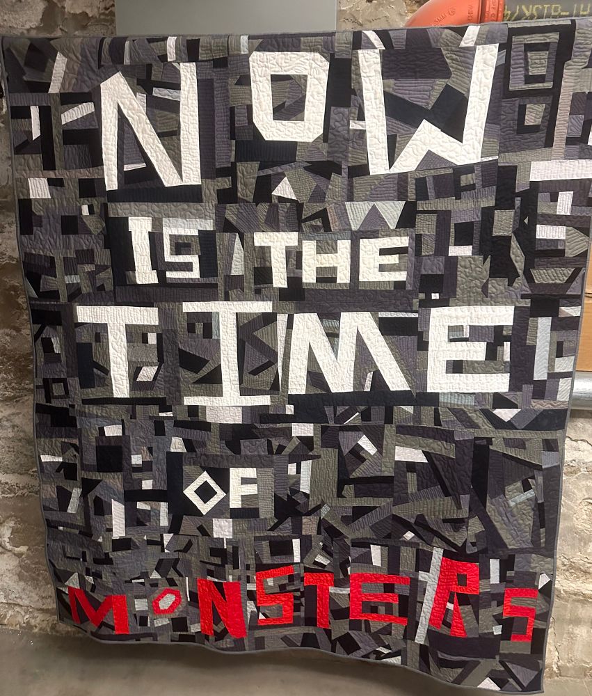 "Now is the time of monsters" is the quote this quilt's patchwork created in red and white on a crazy quilt background in shades of grey. 