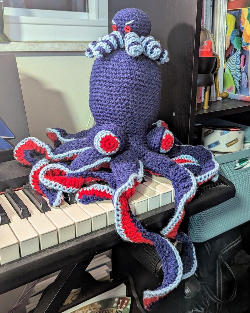 a large crochet octopus in the seattle kraken colors, sitting on a keyboard. atop his head is a tiny crochet octopus in the same colors.