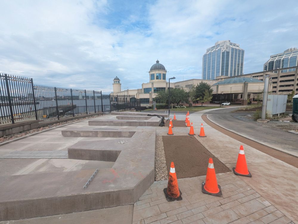 A small park under construction next to a sidewalk and parallel asphalt bike lane. It has several low, concrete plinths that look somewhat like the letters "W" and "U" from above, meant to resemble the wooden piers of the Halifax waterfront that project into Halifax Harbour. The area is still under construction and there are various orange traffic cones sitting around.