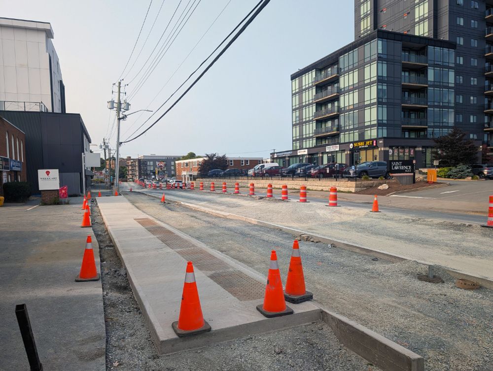 Dutch Village Road in front of the St Lawrence Place apartment building. The road is largely a gravel construction site, but there are disconnected sections of new concrete curbs and sidewalks.