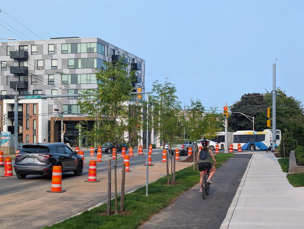 Dutch Village Road, an urban road in Halifax, at the intersection with Joseph Howe Drive. The street is under construction, but this section is nearing completion. A strip of trees and a bike lane have been added between the roadway and sidewalk, which have been completely reconstructed. A woman is riding a bicycle on the bike lane. There's a Halifax Transit bus in the background.