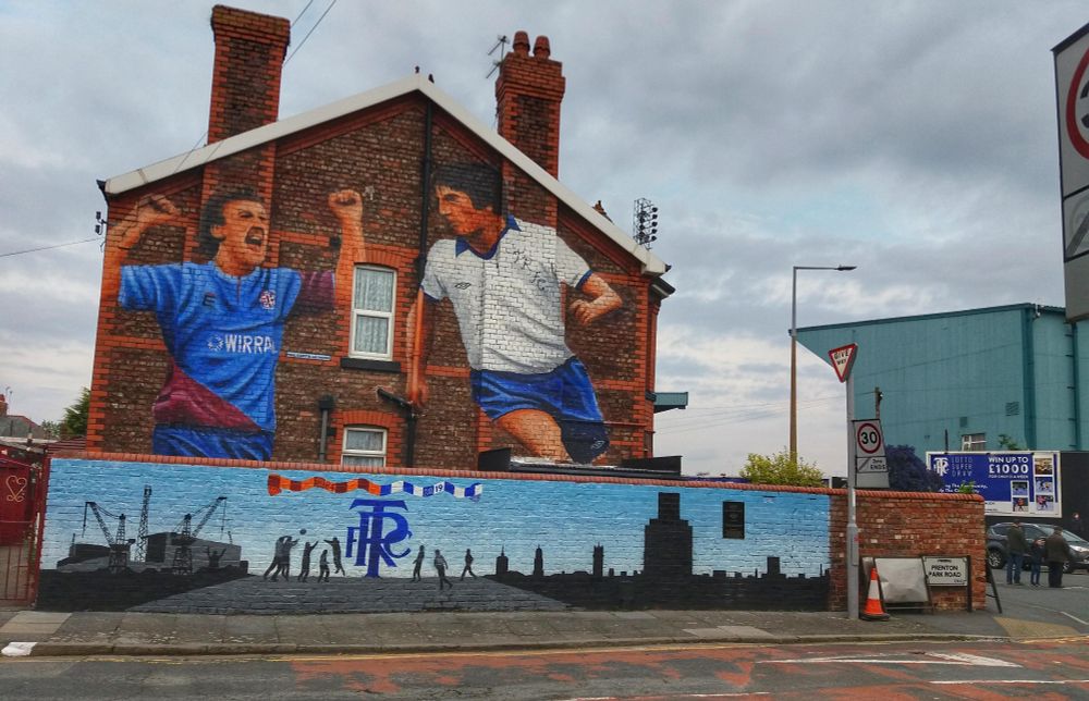 A gable end of a row of terraced houses, covered with a mural depicting two football players in action. The ground can be seen background right. 