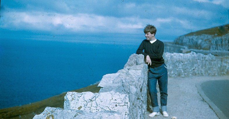 A gawky boy leaning on a stone wall looking out over the sea.