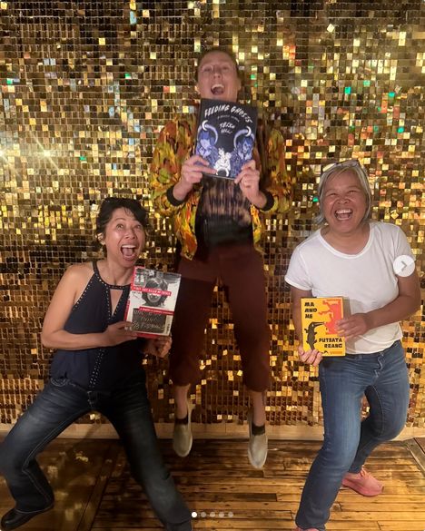 An informal photograph of three women posing in front of a glittery backdrop.  Each is holding a book.