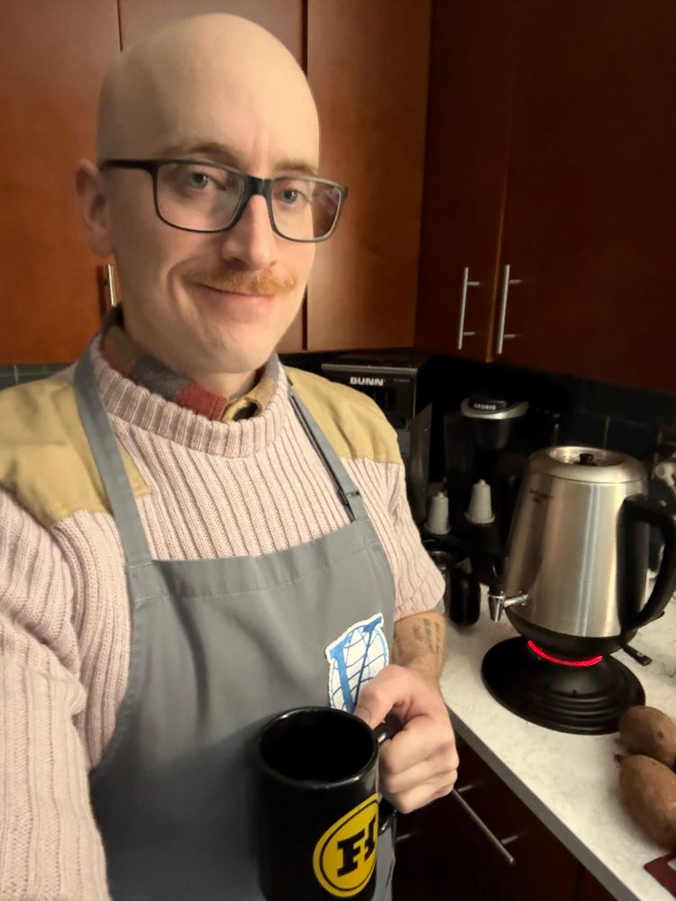 Me holding a cup of coffee in a Funhaus mug as my samovar heats up. I have on a sand colored commando sweater and a grey apron with Venture Industries logo from the Venture Bros