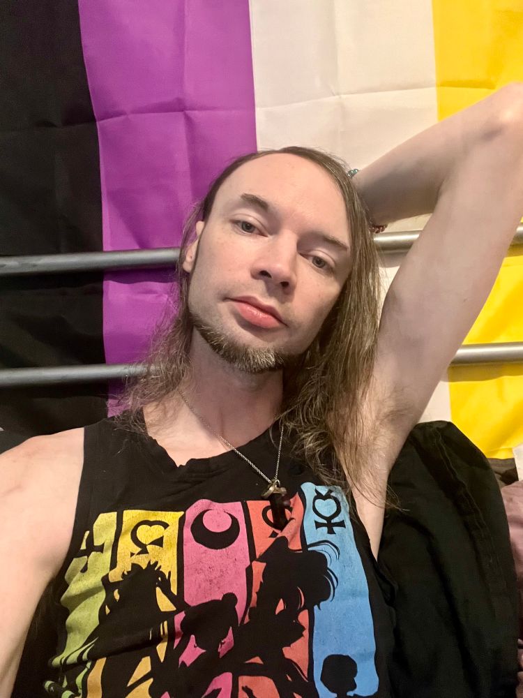 Messy hair boy flashing pit in front of an enby flag. 