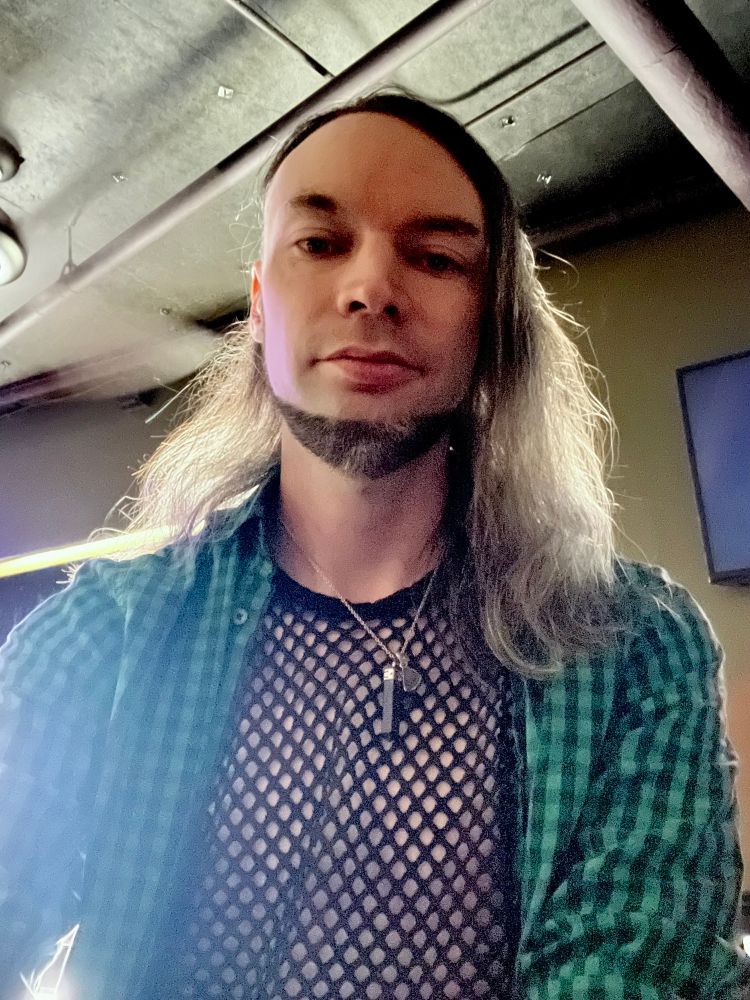 Long hair boy with a short beard in a fishnet top with the sun at his back, sitting in the gay bar. 
