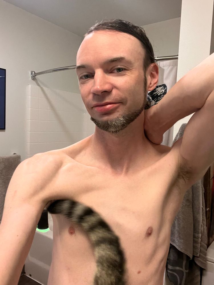 Slim shirtless guy with hair tied back getting photobombed by a cat tail. 