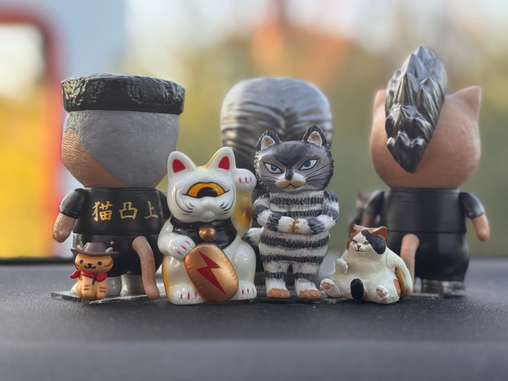 A line up of small cat toys including a tiny cowboy cat from Neko Atsume, a one eyed maneki Neko by Realhead, the cat that lived 1,000,000 times, and a chunky calico cat sitting like a person holding a gold coin under it’s left arm and holding his right paw out as if to tell you to come here. Behind these are three larger soft vinyl cats in outfits with haircuts such as pompadour and Mohawk so they look like Japanese tough guys 