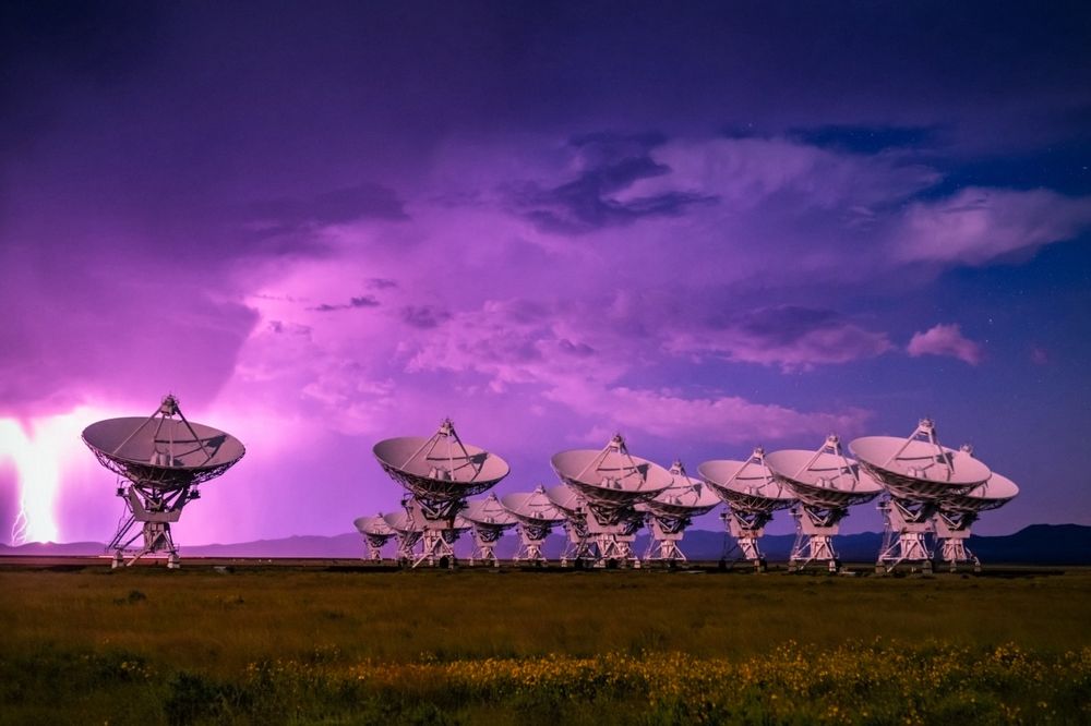 A photo showing an array of radio telescopes pointing in the same direction, acting in synchrony as a larger radio telescope. The sky is dark and a bolt of lightning flashes in the background.