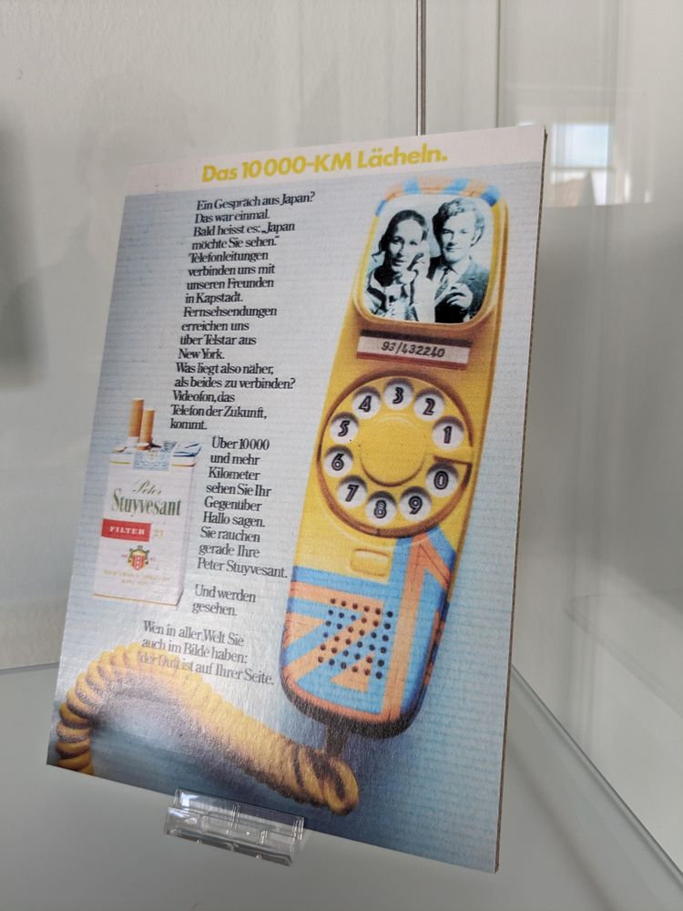 Im Grunde Werbung für Zigaretten.
Neben einer solchen Schachtel ein Bild von einen verkabelten Handteil eines Telefons in orange-blau mit Wählscheibe und kleinem Bildschirm.