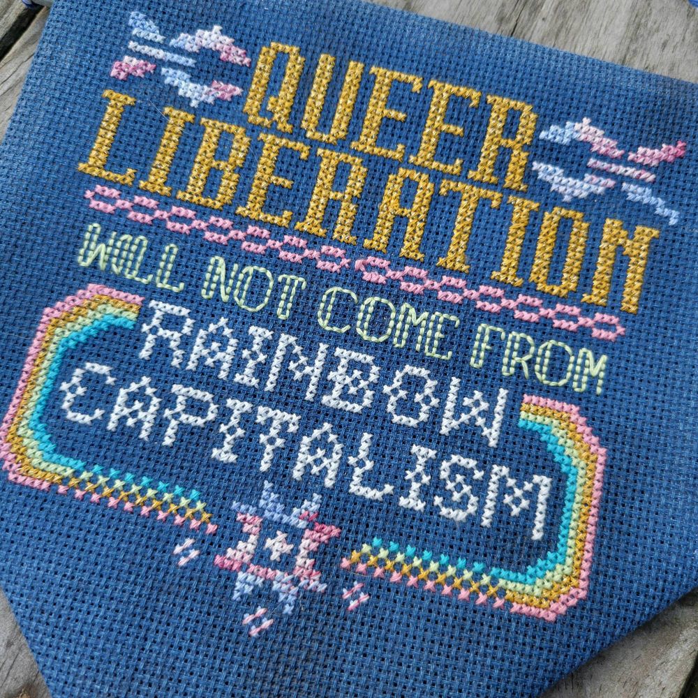 A pennant made of navy blue aida. Brightly colored cross stitching features the words "queer liberation will not come from rainbow capitalism" along broken chain motifs, a rainbow, and a starburst. The starburst & chains are in the colors of the trans flag. 

