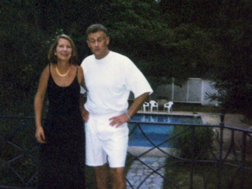 A woman in a black dress and white pearl necklace stands next to a man in white shorts and t-shirt in front of a pool
