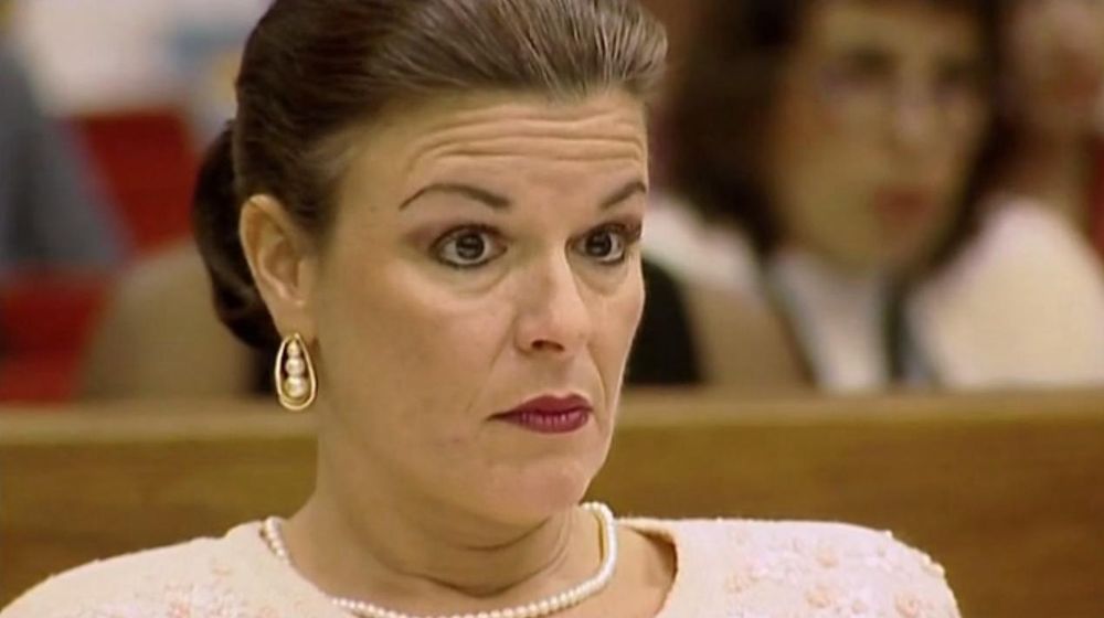 A white woman with dark hair in a bun, wearing a pearl necklace and a white top, looking surprised