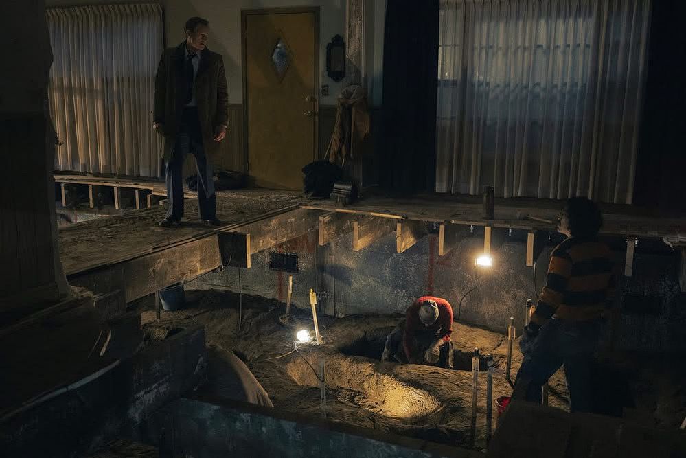 A man in a dark suit looks down at a massive hole in an excavated floor when a person is digging using assistive lighting