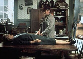 An older white man in an apron and cap stands next to the laid out body of a woman on a table