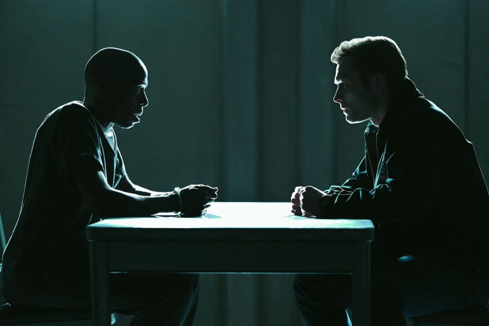 The silhouette of a black man in handcuffs stares at a white man on opposite sides of a prison room table