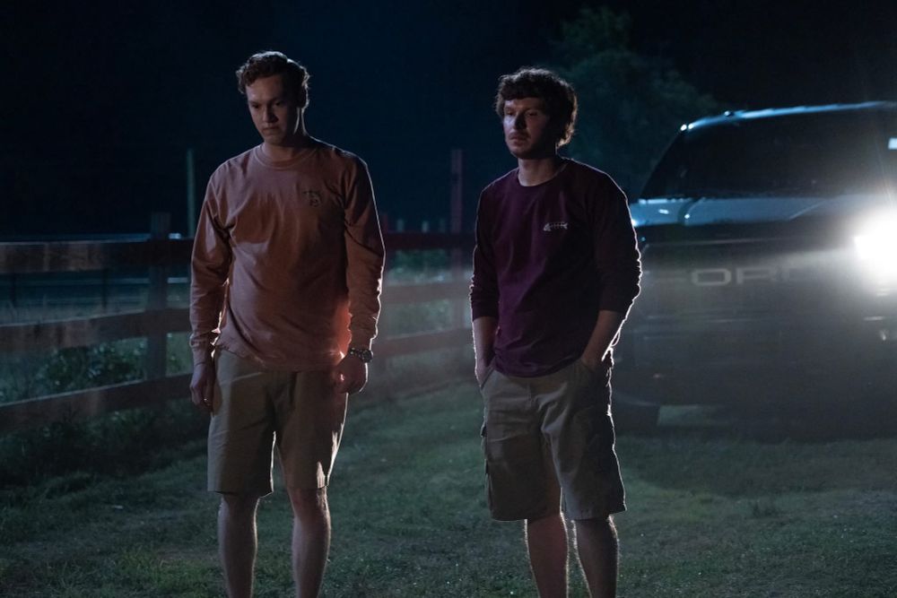 Two redheads (one in a pink shirt; one in maroon) stand in the headlights of a black Ford SUV on a dirt road