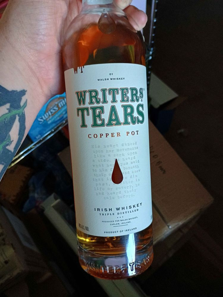 A bottle of amber liquid with a white label and green writing that says Writer's Tears Copper Pot Irish Whiskey, held in a hand with the start of a tattoo visible