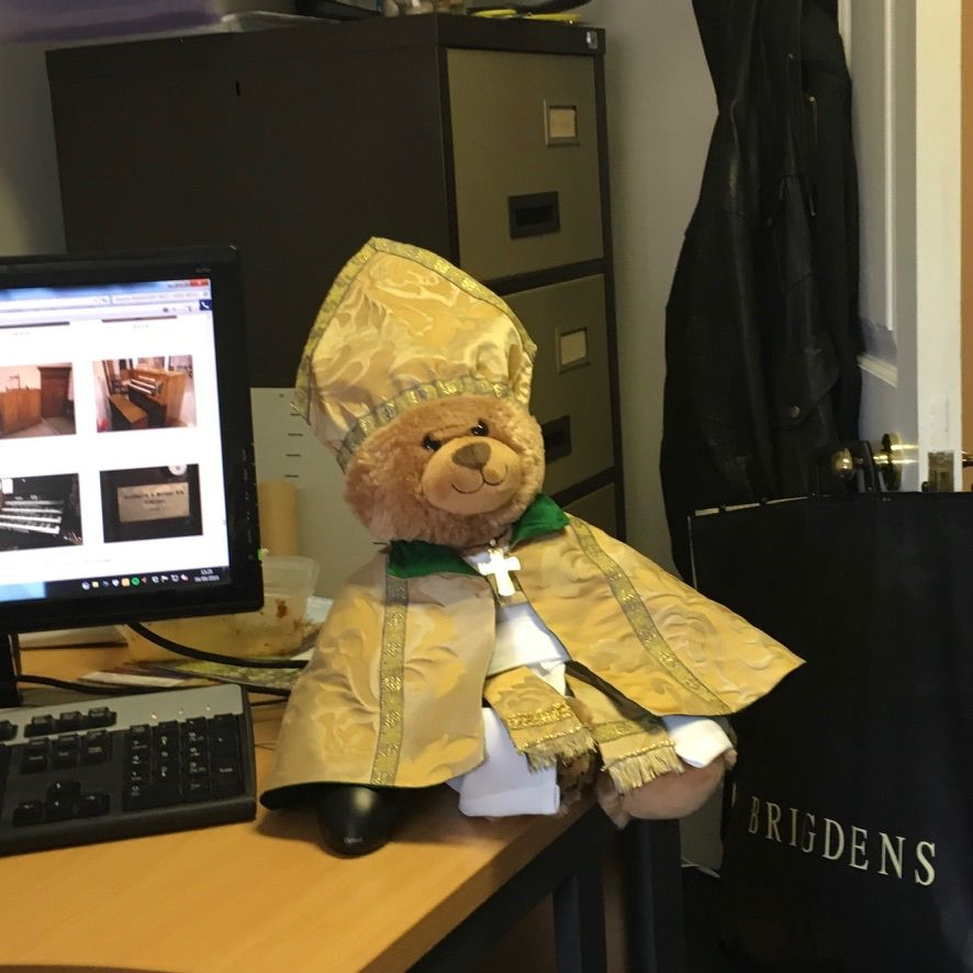left to right: computer screen, desk, teddy bear dressed as bishop in cope and mitre, filing cabinet