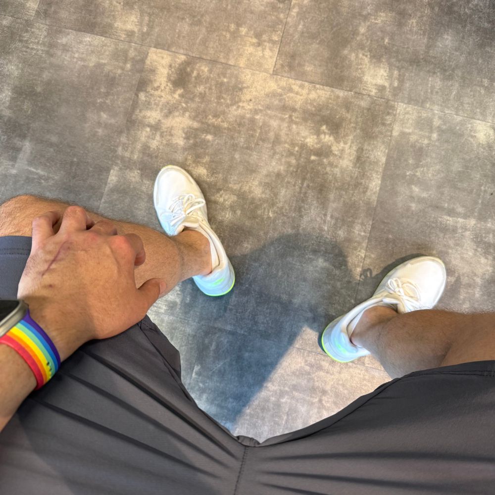 Deutsch:

Eine Person sitzt oder steht leicht breitbeinig auf einem grau gemusterten Boden. Sie trägt weiße Sportschuhe mit einem Hauch von Neonfarbe, eine dunkelgraue #Sporthose und ein #Fitnessarmband in Regenbogenfarben. Die linke Hand ruht auf dem Oberschenkel, sichtbar ist eine Narbe auf dem Handrücken. Das Bild vermittelt den Moment vor oder nach einer sportlichen Aktivität.

Englisch:

A person is sitting or standing with legs slightly apart on a grey patterned floor. They are wearing white athletic shoes with a hint of neon color, dark grey #sports shorts, and a #rainbow-colored fitness wristband. Their left hand rests on their thigh, and a scar is visible on the back of the hand. The image captures a moment before or after a workout session.