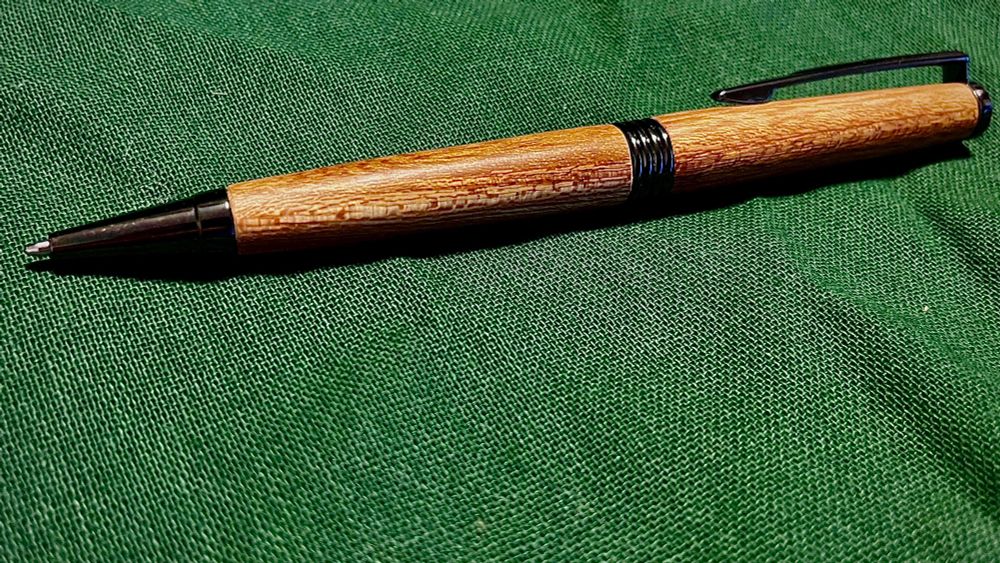 A wooden pen lying on a green fabric background. The wood is orangey brown and lightly patterned. 
It’s fairly unremarkable as far as pens go, but I made it! 