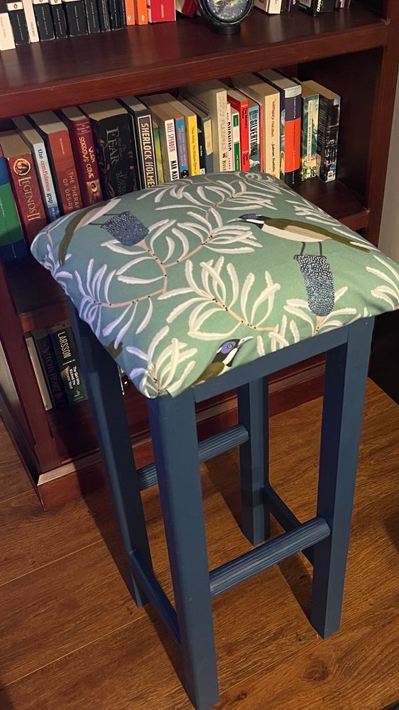 Looking down at the wooden bar stool I made, you can see the dark grey-blue of the painted legs, and the sage green fabric patterned with silver banksia leaves and blue and grey birds.
The stool is positioned in front of a bookshelf with a bunch of book titles visible (including When Sorrows Come by Seanan Maguire, Wise Man’s Fear by Patrick Rothfuss, and Man Made Language by Dr Dale Spender).