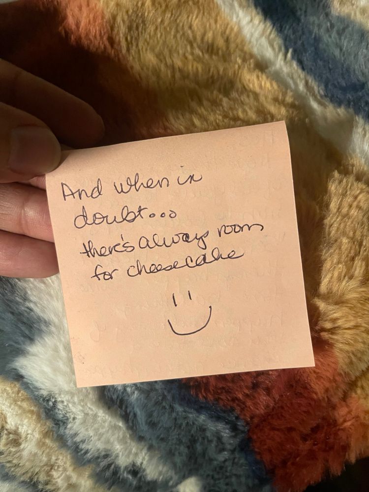 The back of the same peach-colored sticky note, photographed against the same soft blanket. The handwriting reads:
“And when in doubt… there’s always room for cheesecake.”
A small smiley face is drawn underneath.
