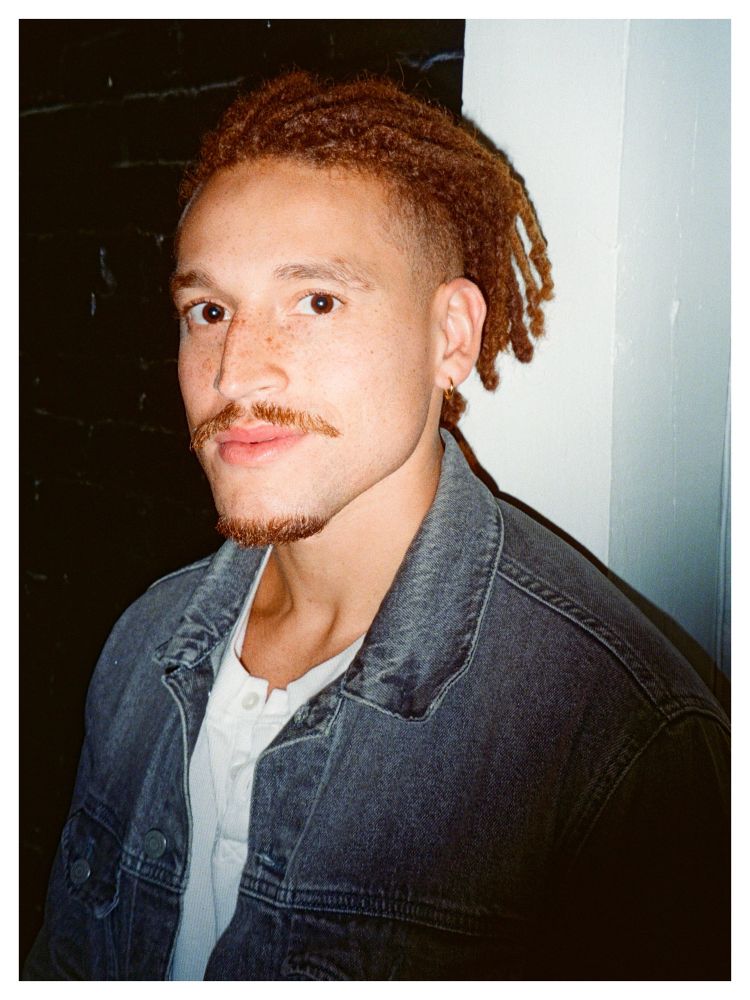 A person with light brown locs pulled back wears a denim jacket over a white shirt. They stand beside a dark wall, looking slightly past the camera, illuminated by bright flash.