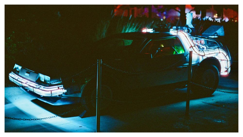 DeLorean time machine lit up at night, glowing blue and white under neon lights with red highlights in the background.