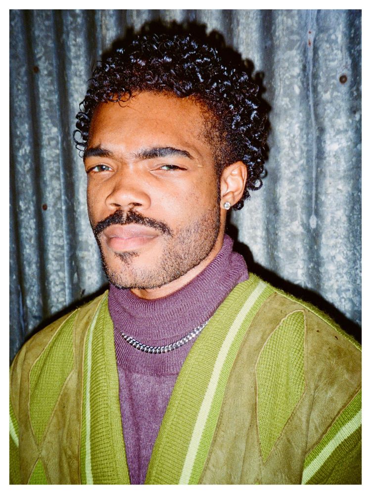 A person with short, tightly curled hair and a trimmed beard wears a green and tan sweater layered over a purple turtleneck. They stand in front of a corrugated metal wall, giving a subtle squint under direct flash.