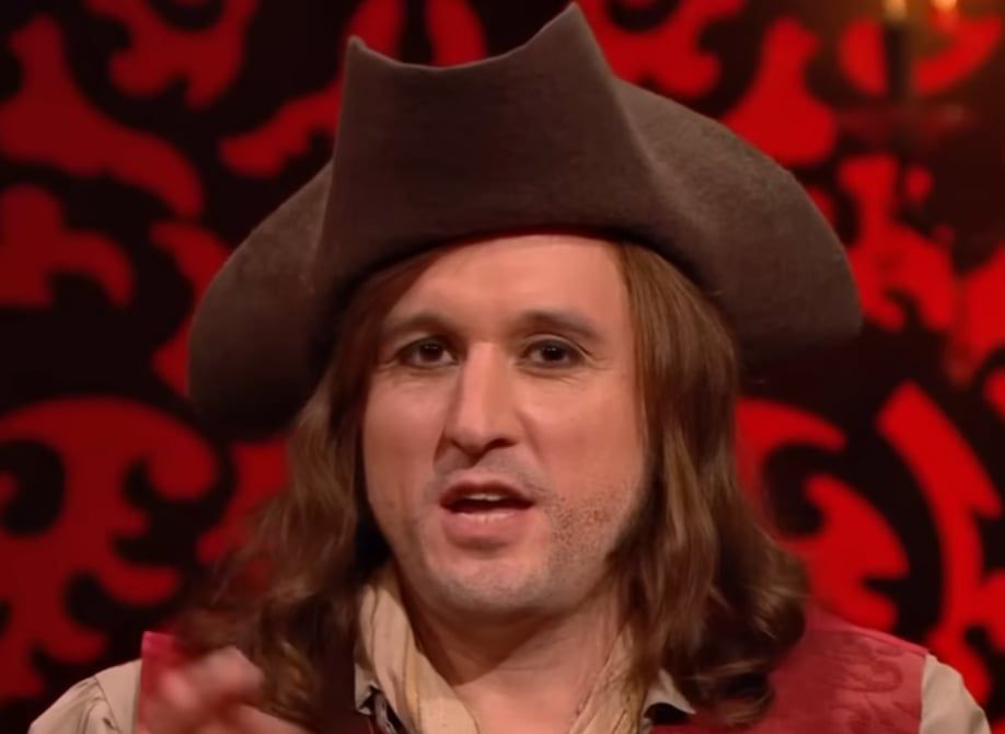 A man sits candidly in front of a red and black background. He is wearing a three-point pirate hat and a long-haired brown wig. This is British comedian Phil Ellis. 