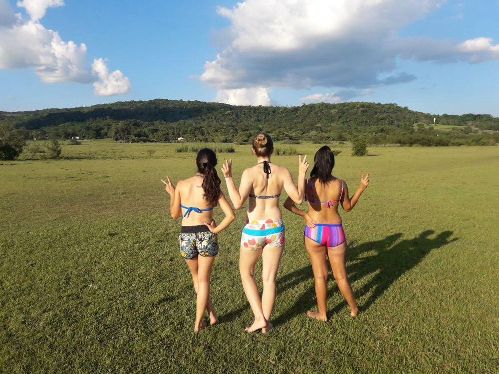 Three girls stand with their backs to the camera, posing for a photo in swimsuits. The middle girl has her blonde hair in a bun and the other two girls have darker hair in ponytails. The background is of a cerro nearby Quyquyho, Paraguay.