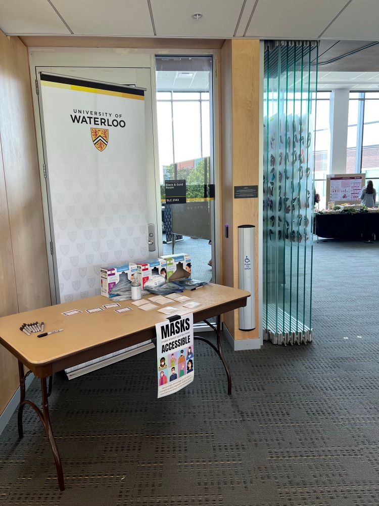 Entrance to the symposium room. Dark grey carpet and opened glass doors.

A brown table with boxes of N95 respirator mask, a sign that says masks make shared spaces more accessible taped to the front, and a UWaterloo vertical banner behind the table.