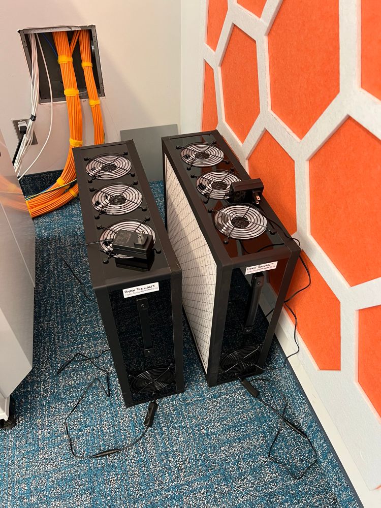 Two black PC fan air cleaners sitting in the corner of the room on blue carpet in front of an orange wall (they are in storage for use at the symposium event)