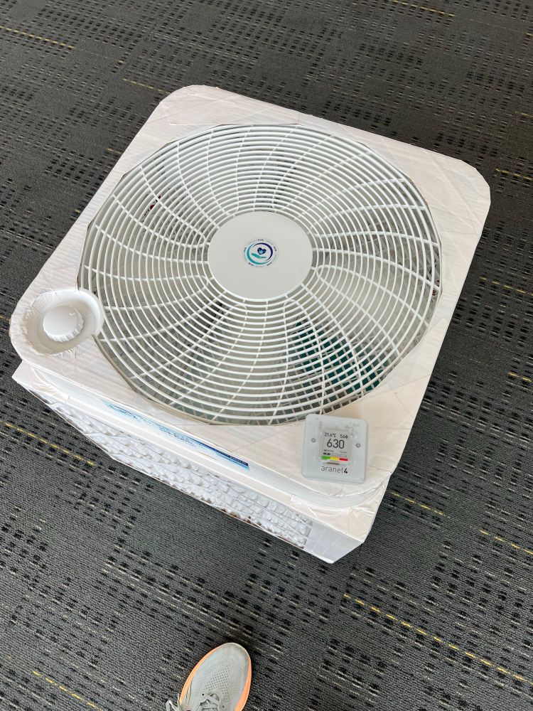 A close up photo of a 20 inch box fan attached to four MERV 13 filters. An Aranet monitor sitting on top of it shows 630 ppm. You can see a blue shoe in the image on the dark grey floor.