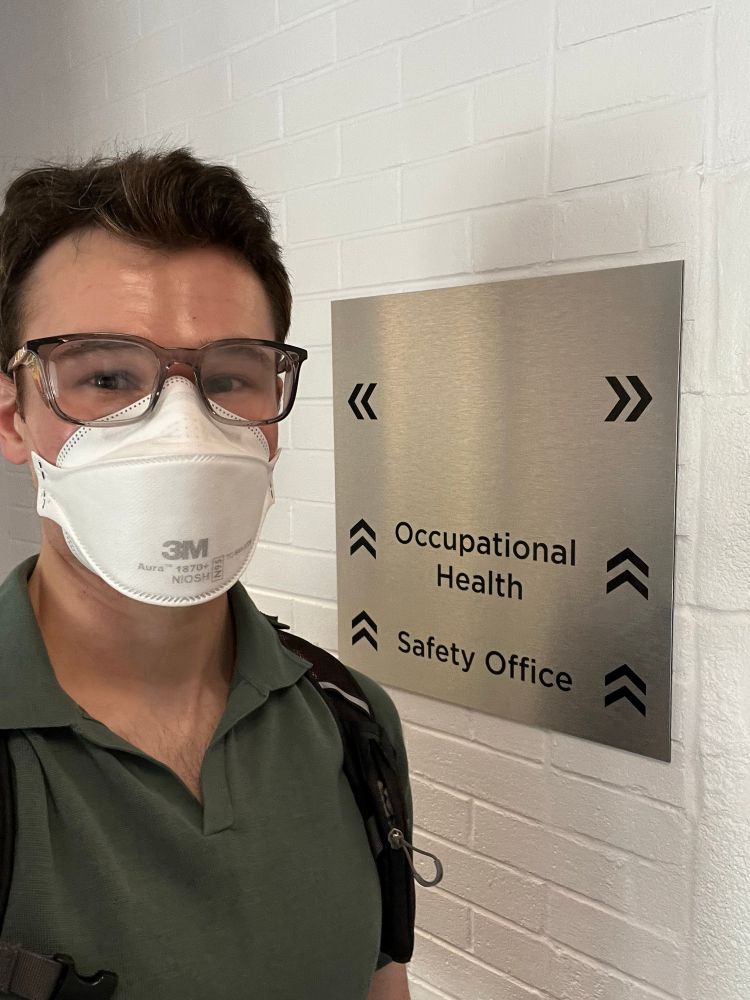White man with short brown hair, stylish clear safety glasses, a white 3M n95 mask,  dark green collared shirt, standing in front of a white brick wall with a silver sign that says occupational health and safety office with chevron directional arrows.