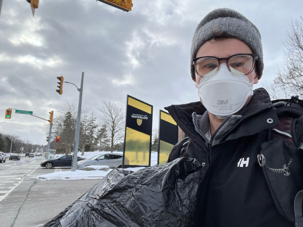 A selfie phot of a white male with glasses and a grey hat, wearing a black winter coat standing on the corner of a road. A black and yellow university of Waterloo sign can be seen in the background along with cars and traffic lights. He is carrying a black garbage bag.
